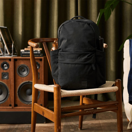 A black backpack sits on a wooden chair beside a speaker system and potted plant. It features multiple compartments and a logo.