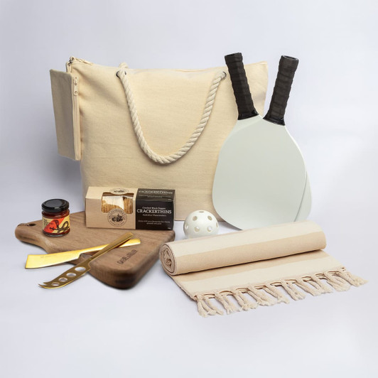A cream tote bag, wooden board, drink jar, cheese knife, paddles, ball, picnic blanket, and gourmet snacks.
