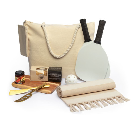 A picnic set featuring a beige tote bag, paddle bats, a ball, a cutting board, a knife, and a blanket.