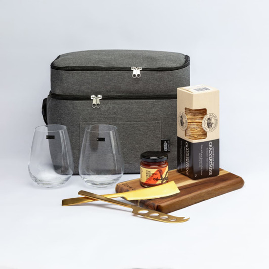 A grey cooler bag with two glass tumblers, a wooden cutting board, cheese knife, jar of chutney, and crackers.