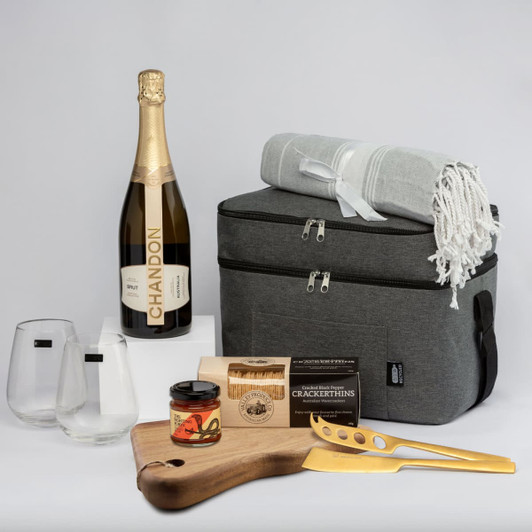 A grey cooler bag with a blanket, two glasses, a bottle of Chandon, a cheese board, and gourmet products inside.