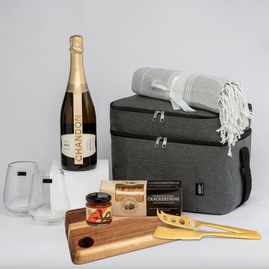 A grey cooler bag, bubbly drink bottle, two stylish glasses, a wooden serving board, a jar, and a blanket.
