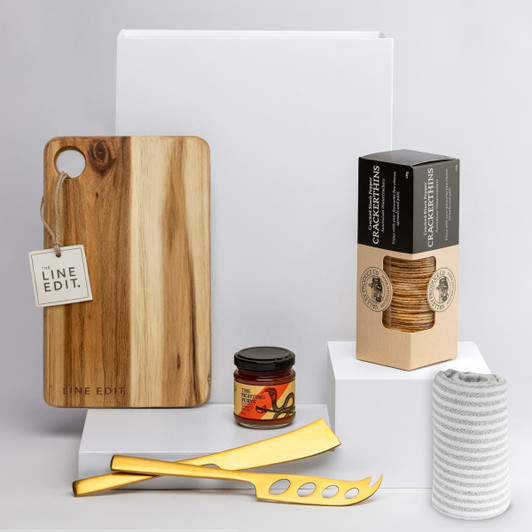 A wooden cheese board with a tag, gold cheese knives, a jar, a box of crackers, and a coiled cloth.