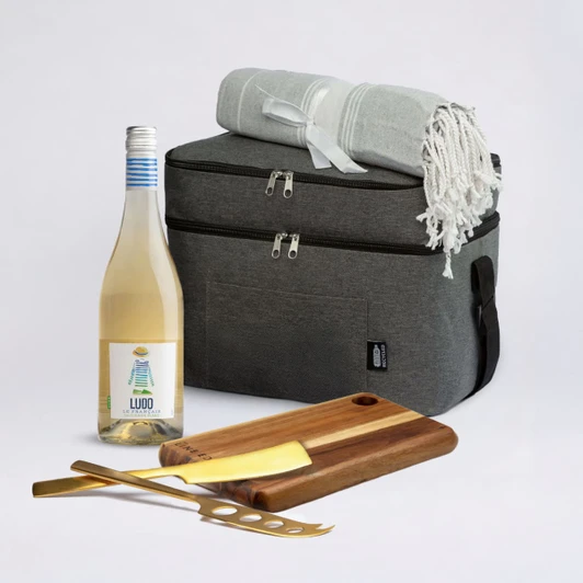 A cooler bag in grey with a cream blanket, a bottle of wine, and a wooden cheese board with utensils.