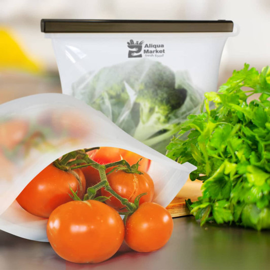 A silicone reusable storage pouch in transparent white containing broccoli, with fresh tomatoes and parsley beside it.