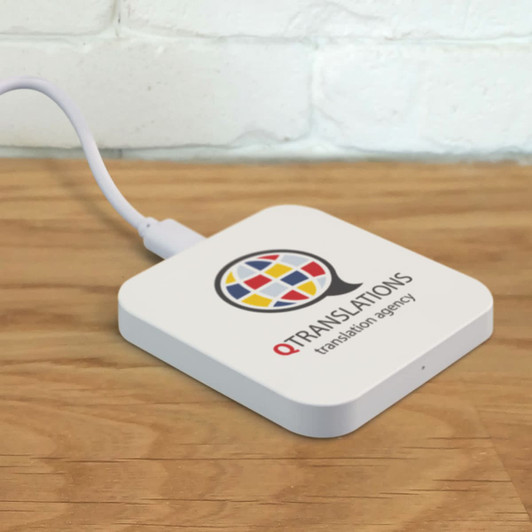 A square white wireless charger with a branding logo on top, connected by a cable on a wooden surface.