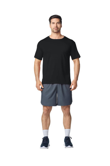 A man stands wearing a black Gildan Performance adult T-shirt and grey shorts, with sports shoes and socks.