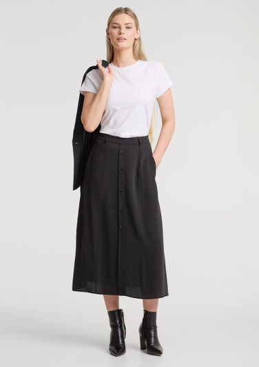 A woman stands wearing a white t-shirt and a black midi skirt, paired with ankle boots. The skirt features buttons and has a logo.