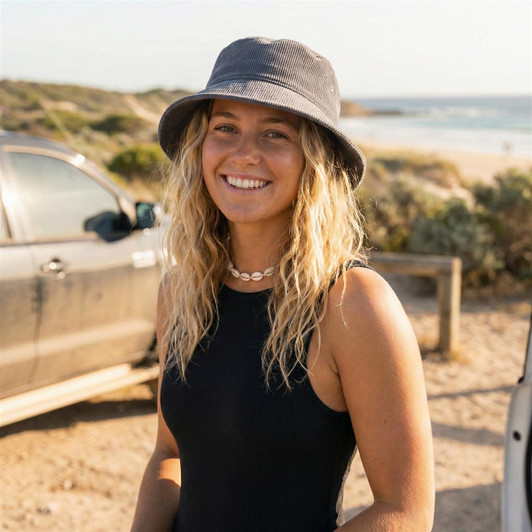 A woman stands by a beach wearing a grey cord bucket hat, smiling with wavy blonde hair.