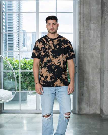 A man wearing a black and tan bleach-dyed T-shirt and distressed denim jeans, standing in a modern interior.