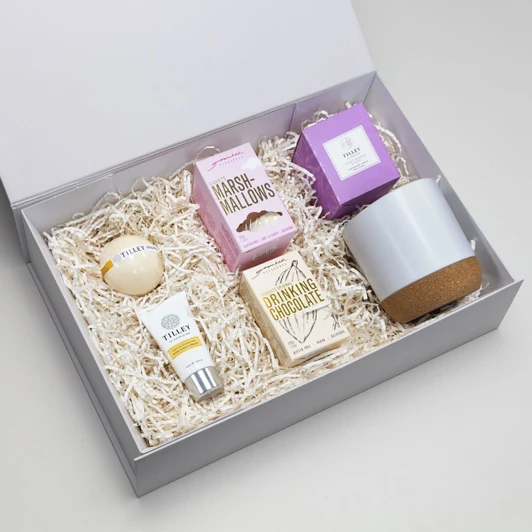 A gift box containing a grey pottery cup, drinking chocolate, marshmallows, and assorted skincare items.