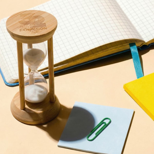 A bamboo hourglass with sand sits beside a notebook, a yellow notepad, and a blue sticky note with a paperclip.