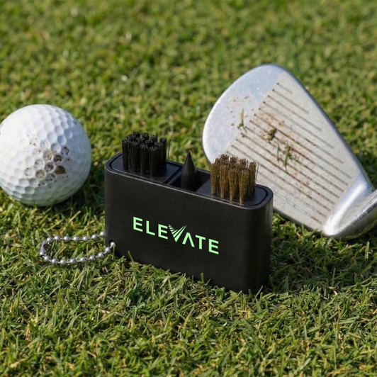 A black golf club cleaning tool with brushes sits on grass next to a golf ball and a club with dirt on it.