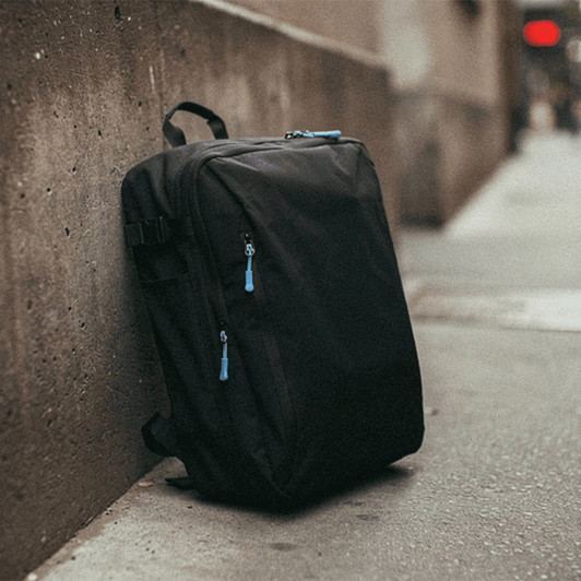 A black travel backpack with blue zipper pulls leaning against a concrete wall. It features multiple compartments.