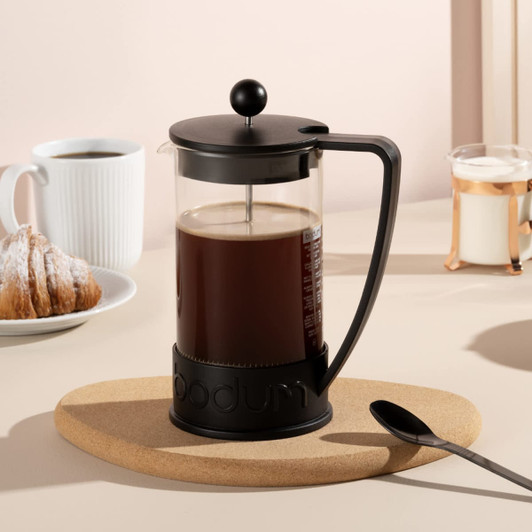 A Bodum Brazil French Press in black, filled with coffee, set on a cork mat, with a spoon and pastries in the background.