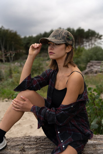 A woman wearing a camouflage cap poses outdoors, with a checked shirt and a natural backdrop of trees and sand.