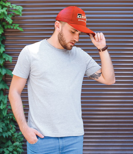 A man wearing a grey t-shirt and a red cap stands against a textured background with greenery. The cap features a logo.