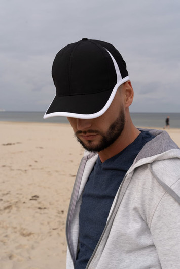 A black cap with white trim worn by a man on a beach, wearing a grey hoodie and a blue T-shirt.