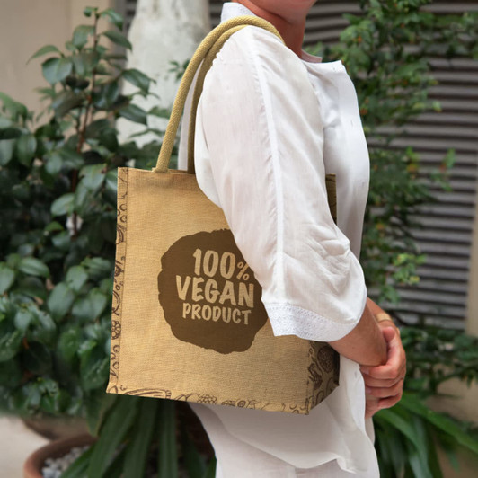 A jute tote bag in brown, featuring a logo, held by a person wearing a white shirt, against a greenery backdrop.
