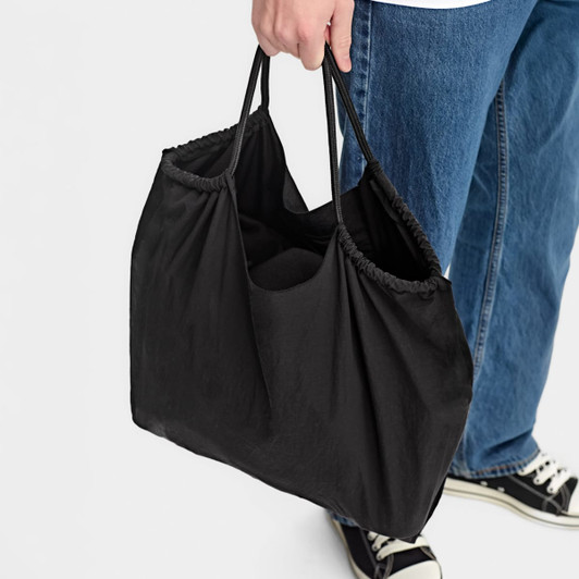 A black nylon shopper bag with two handles, being held by a person wearing jeans and sneakers.