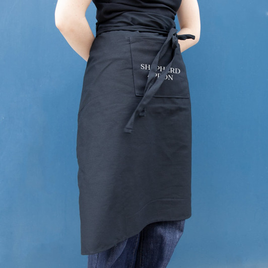 A black long waist apron with a side pocket, featuring a logo on the front.