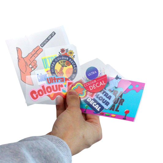A hand holds several Ultra Colour Decal Sheets featuring various colourful designs and a logo.