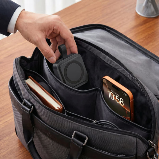A black magnetic wireless charging station is placed inside a dark bag alongside a phone and a notebook.