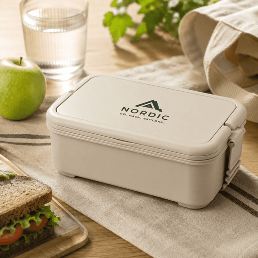 A beige lunchbox with a logo, placed on a table beside a sandwich, an apple, a glass of water, and a cloth.