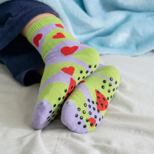 A pair of green and lilac socks with heart patterns and grip dots on the soles, resting on a bed.