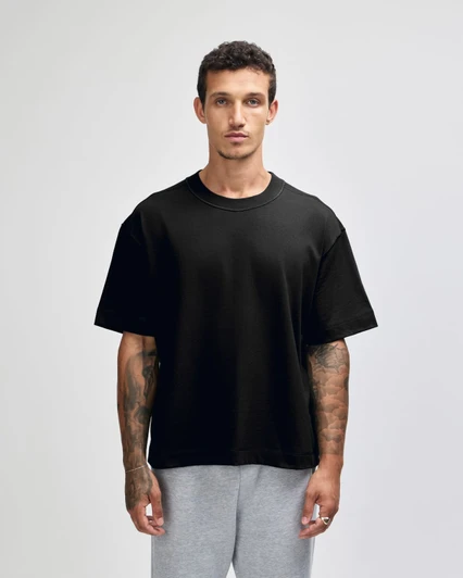 A black unisex heavyweight cotton boxy t-shirt displayed on a model with tattoos, paired with grey trousers.
