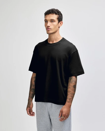 A black unisex boxy t-shirt made of heavyweight cotton, displayed on a model with tattoos, paired with light grey pants.