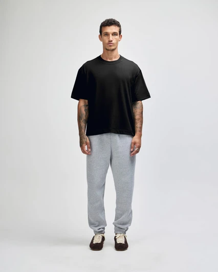 A man stands wearing a black heavyweight cotton unisex boxy t-shirt and grey sweatpants. The image features a logo on the t-shirt.