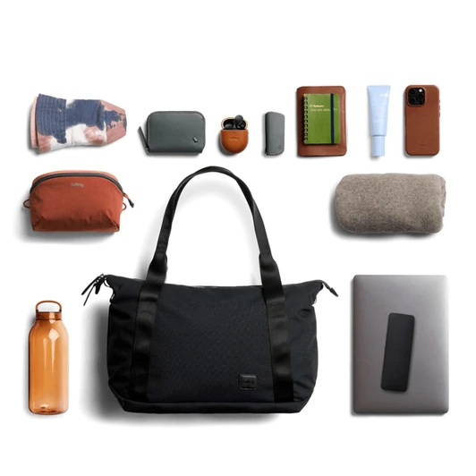 A black tote bag surrounded by various accessories including a drink bottle, pouches, a notebook, and a phone.