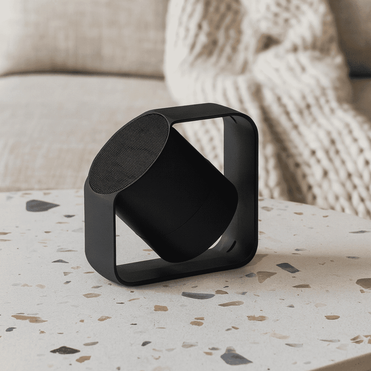 A black wireless speaker with a unique design sits on a speckled table, surrounded by a soft, textured background.