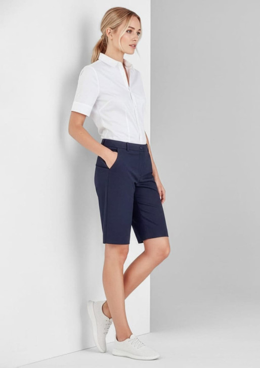 A woman wearing navy women's comfort waist cargo shorts with a white shirt and white shoes, standing against a grey wall.