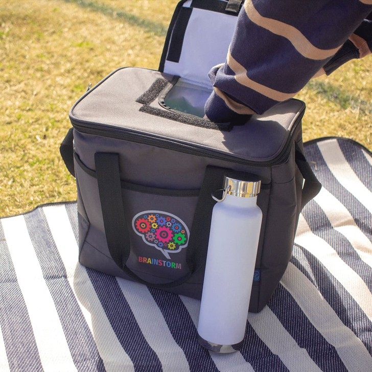 A grey cooler bag with a top opening and handles, featuring a logo, alongside a white drink bottle on a striped blanket.