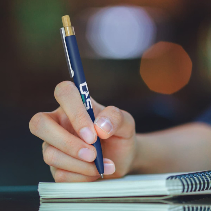 A navy metal pen with a wooden accent held in a hand, poised over a notepad. Features a logo.