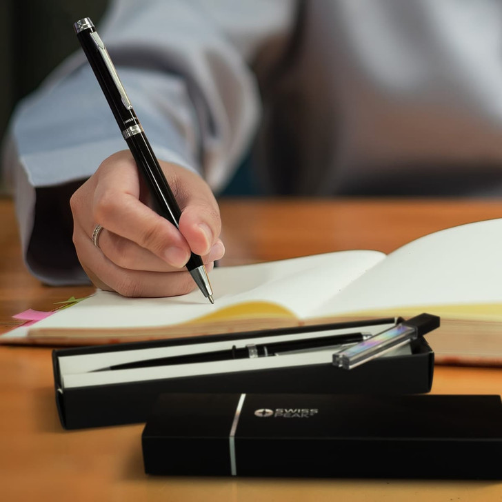 A black pen and pencil set in a sleek box, with a hand writing in a notebook. The set includes a branded logo.
