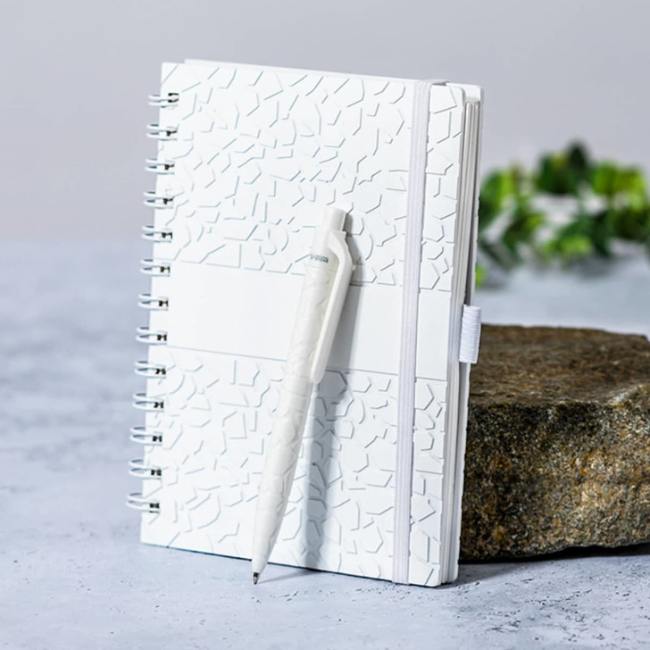 A white spiral-bound notebook with a textured cover, accompanied by a matching pen, set against a natural stone surface.