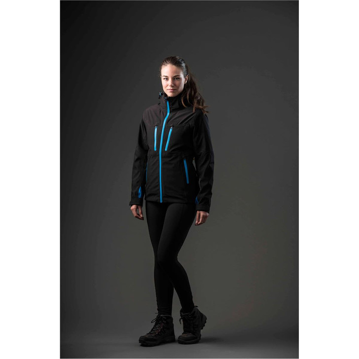 A woman wearing a black jacket with blue accents, paired with leggings and boots, standing against a dark background.