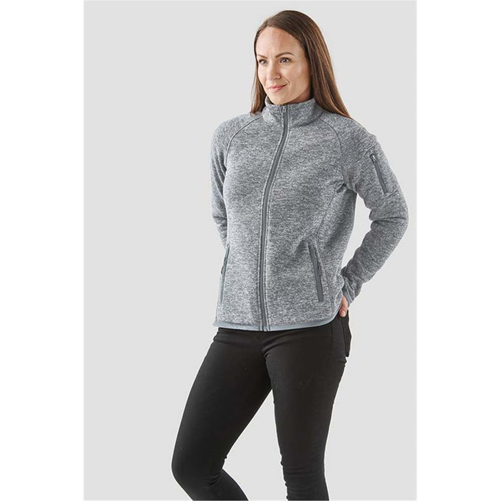 A woman wearing a grey full zip fleece jacket stands with her hands in the pockets, featuring a branding logo.