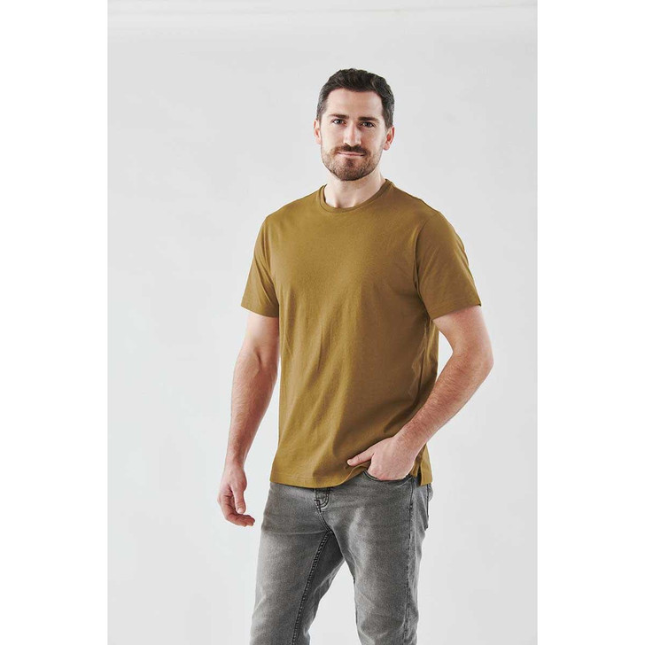 A man wearing a mustard-coloured short sleeve tee, standing against a plain background. The shirt has a logo.