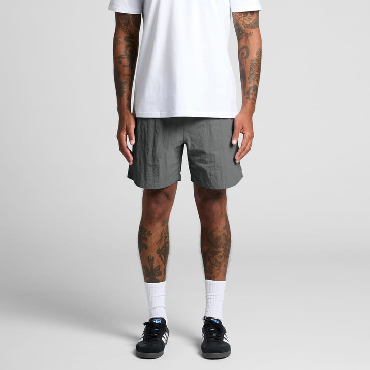 A person stands wearing grey swim shorts, paired with a white t-shirt and black sneakers. The shorts feature a logo.