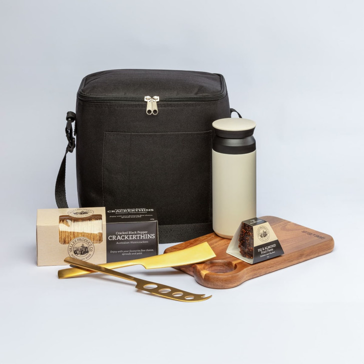 A black cooler bag, a cream drink bottle, a wooden serving board, gourmet snacks, and a gold cheese knife.
