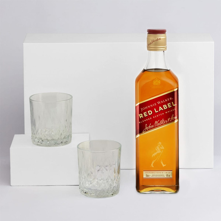 A drink bottle of Johnnie Walker Red Label whisky alongside two crystal tumblers on white podiums.