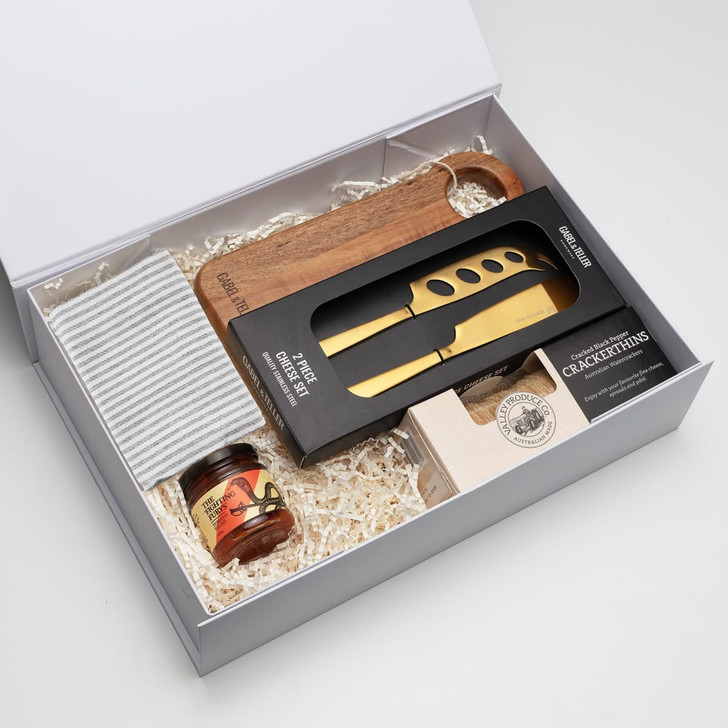 A gift box containing a wooden cheese board, a gold cheese knife, honey, and crackers, arranged on straw.