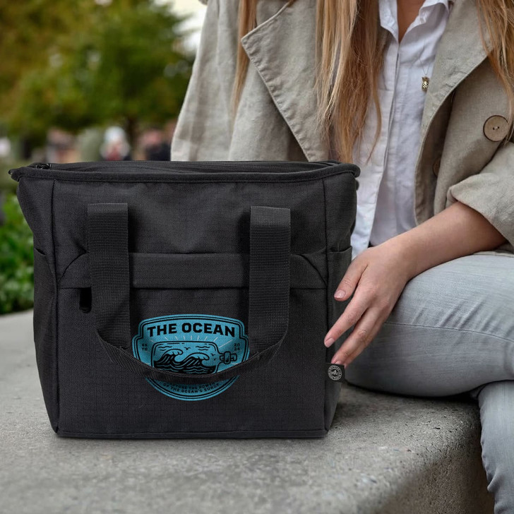 A black cooler bag with a logo sits beside a person wearing a light coat and jeans.