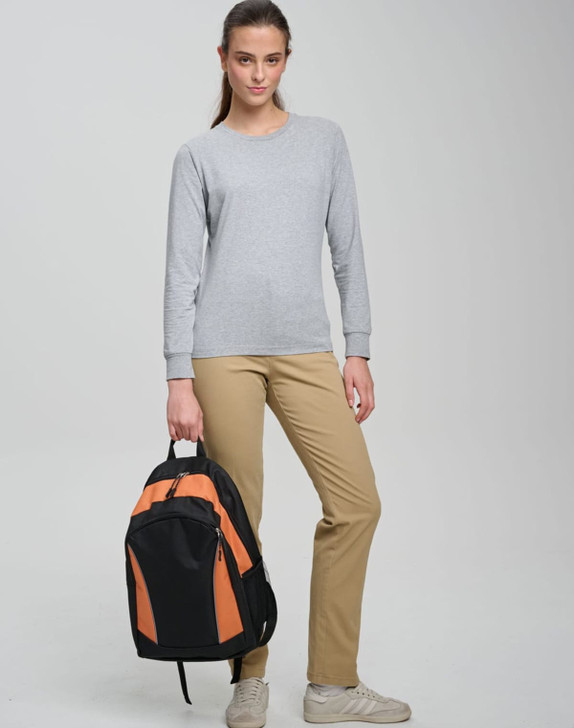 A young woman stands with an orange and black backpack, wearing a grey long-sleeve top and beige trousers.