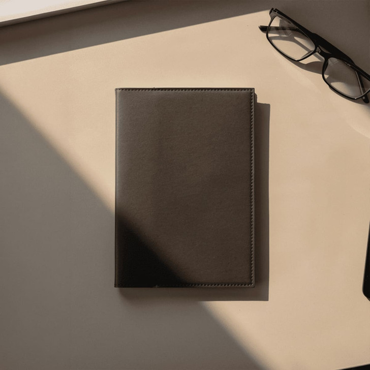 A dark brown leather refillable journal lies on a beige surface, accompanied by a pair of glasses.