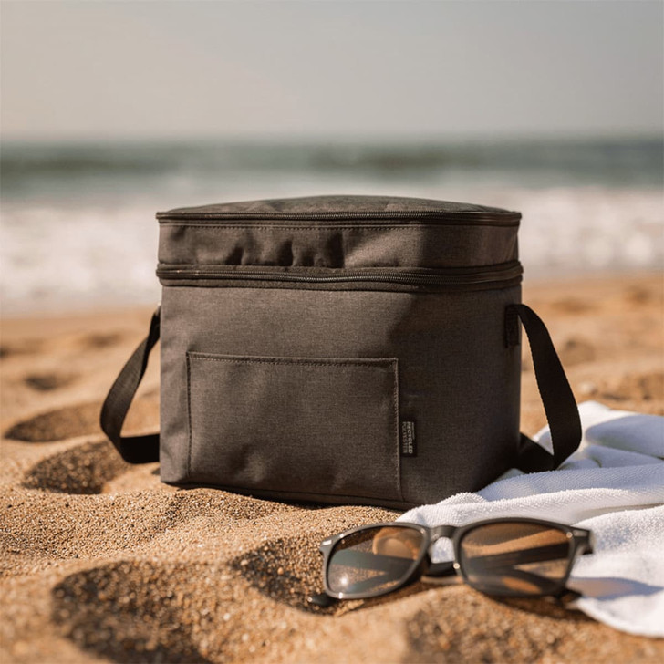 A grey double compartment cooler bag sits on sandy beach alongside sunglasses and a towel, with ocean waves in the background.